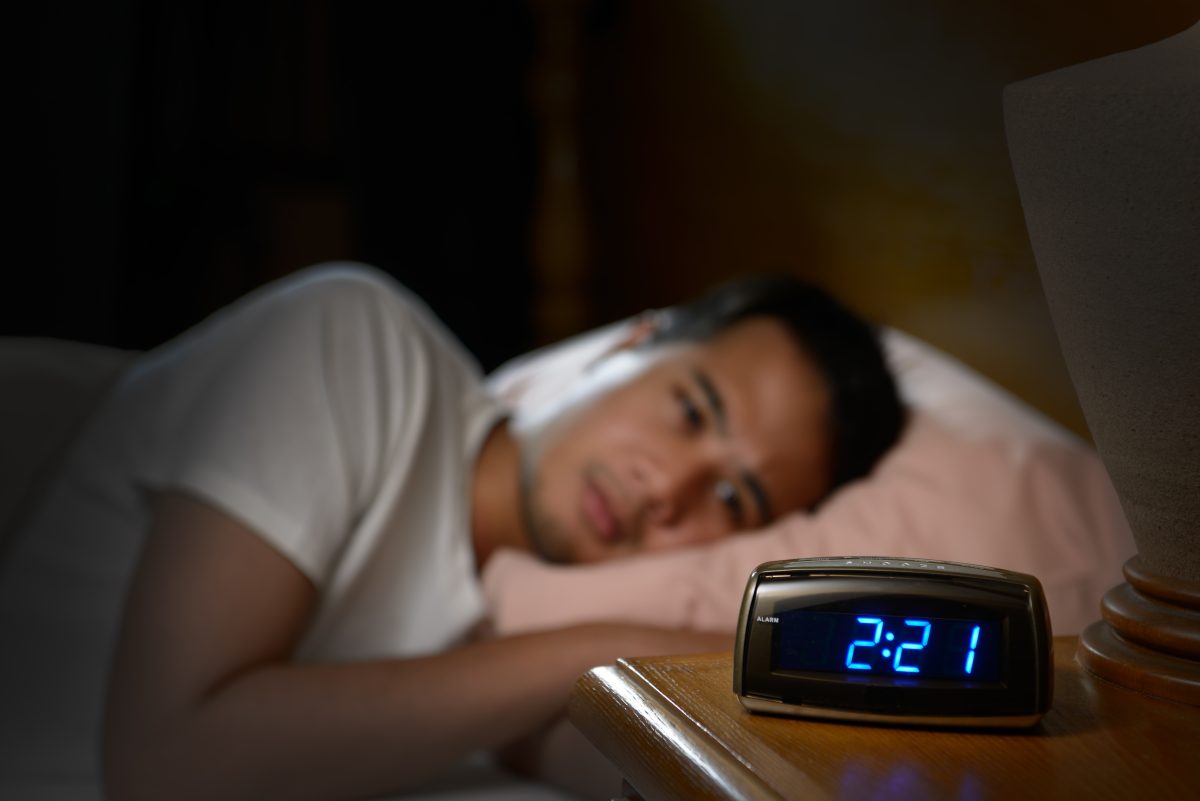 Man lies awake in bed looking at a clock showing 2:21