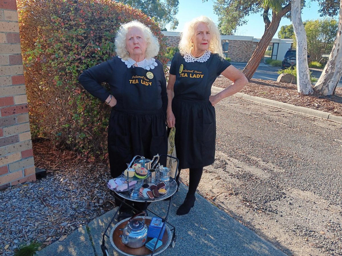 Two white haired women with Tea Lady uniforms standing outdoors in front of a tray of tea cups.