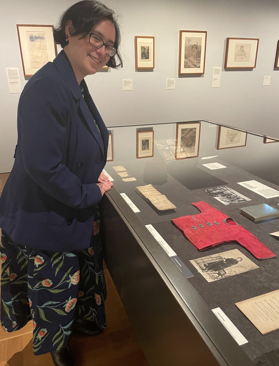 Woman wearing glasses in front of a cabinet displaying papers and a tiny red jacket.