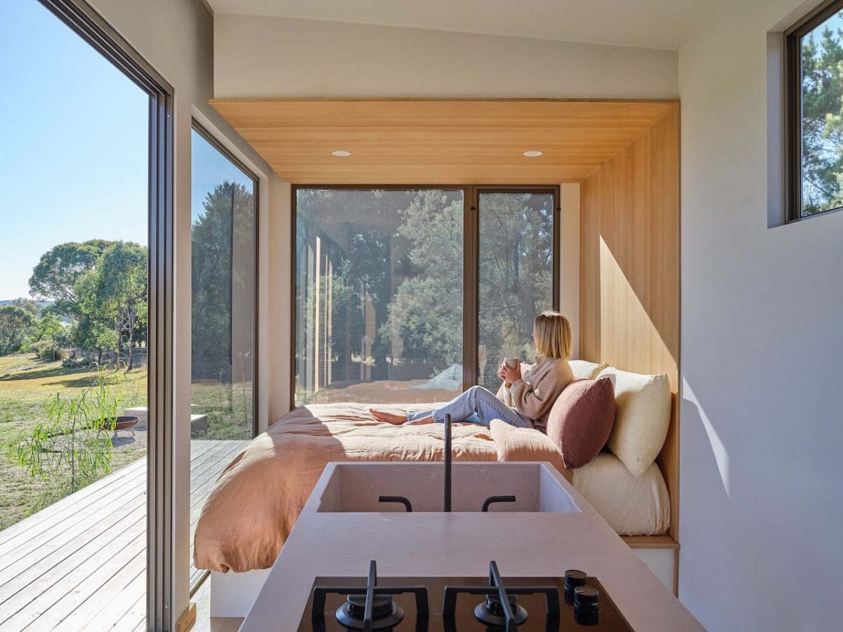 Blonde woman holds a mug while sitting on a bed in a tiny house. Sunlight streams into the room