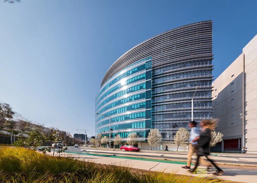 A glass-paned building from the outside with cars driving and people walking on the street out front.