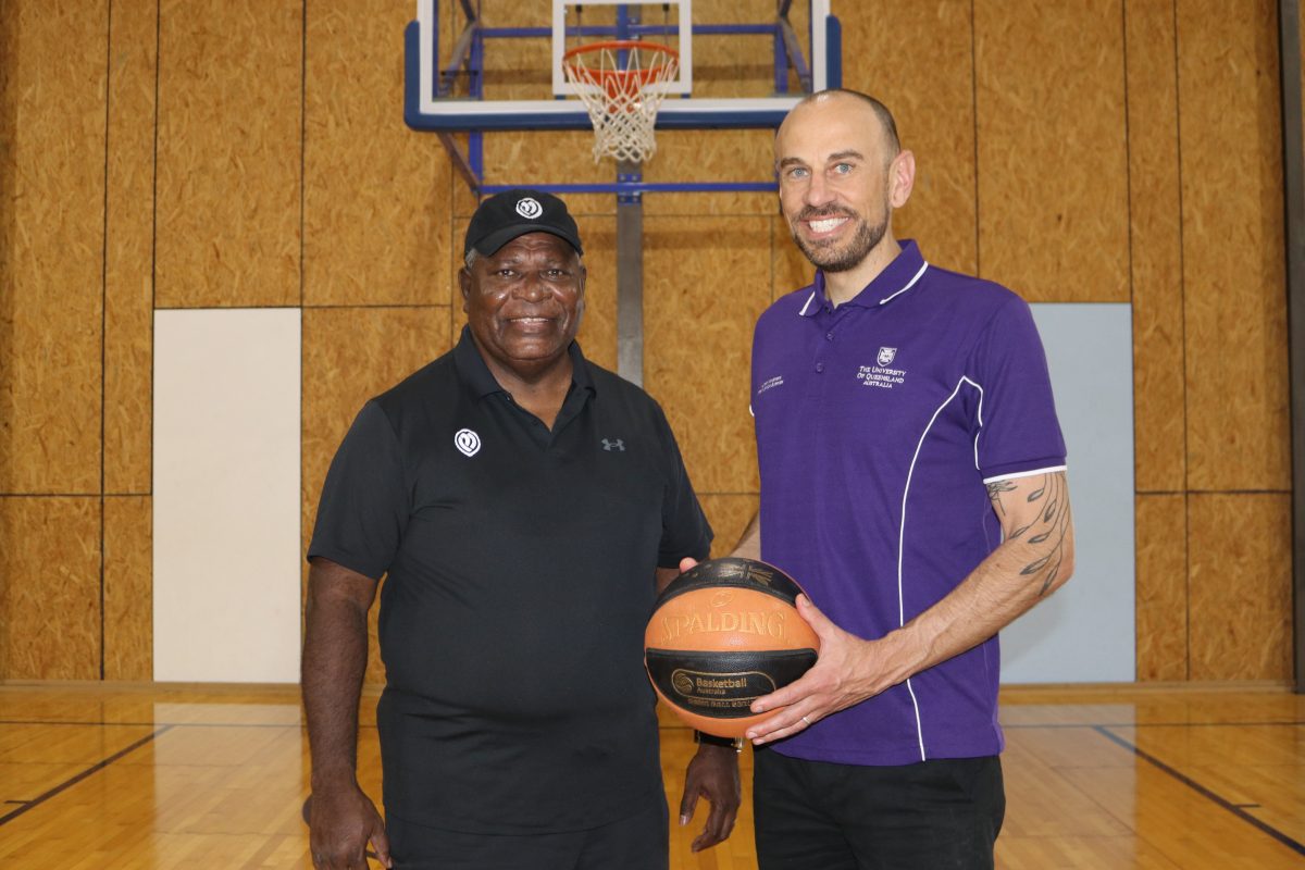 Uncle Benny Mills, wearing a black cap and black polo shirt, is smiling standing next to Dr Wheeler, wearing a royal blue UQ polo shirt and holding out a basketball.