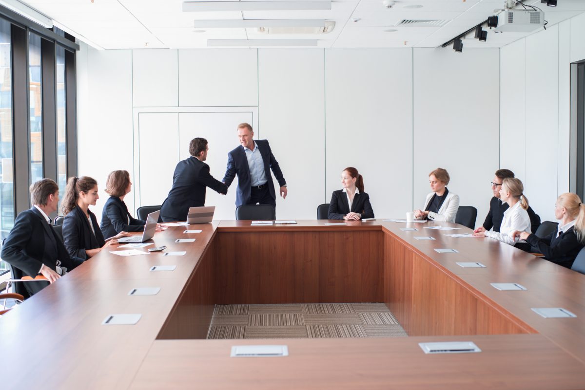 A big meeting in progress, with two men shaking hands as other people look on