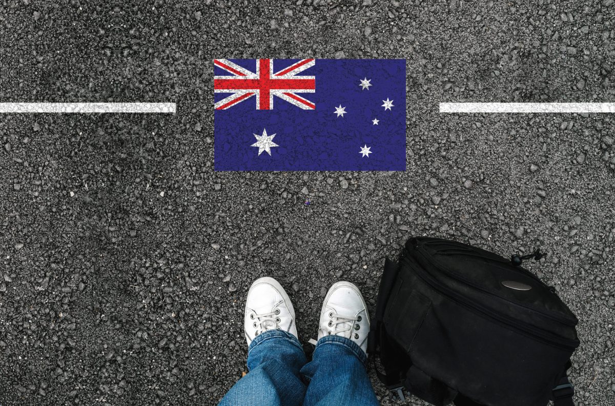 A man with shoes and backpack is standing on asphalt next to flag of Australia and border. 