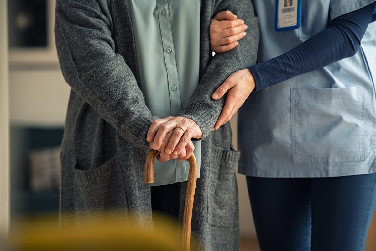 Elderly woman using walking cane at nursing home with nurse holding arm for support