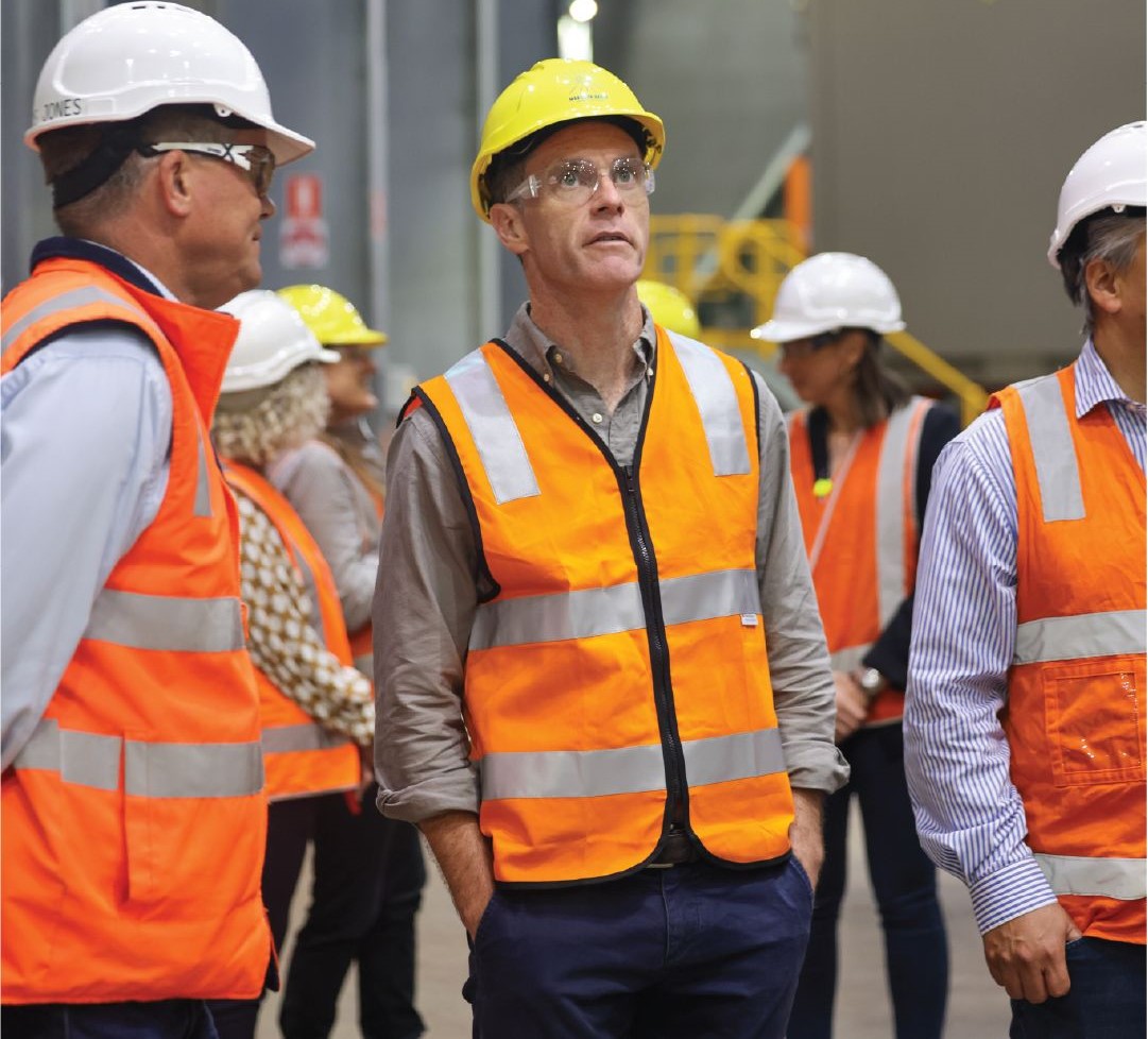 Chris Minns in a high-vis jacket and hardhat, surrounded by similarly dressed individuals in a warehouse.