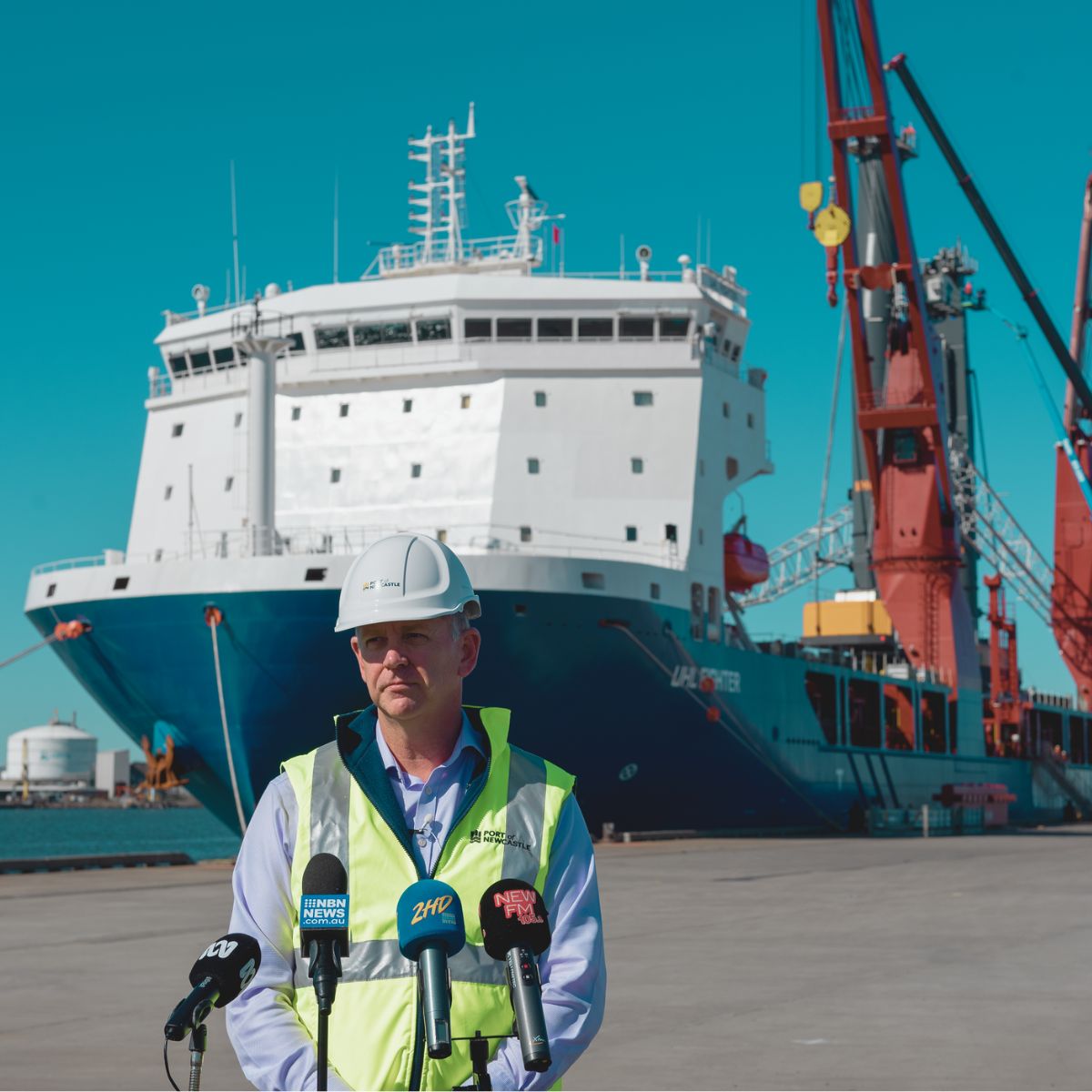 Craig Carmody in a hard-hat and high-vis vest speaking into mics while standing in front of a ship in the dock.