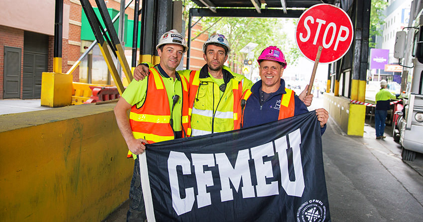 Three construction workers in high-vis holding a CFMEU flag on a street in Melbourne