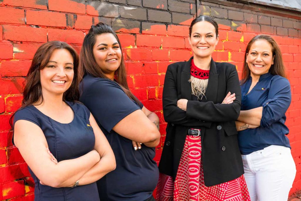 four smiling women standing next to a red brick wall