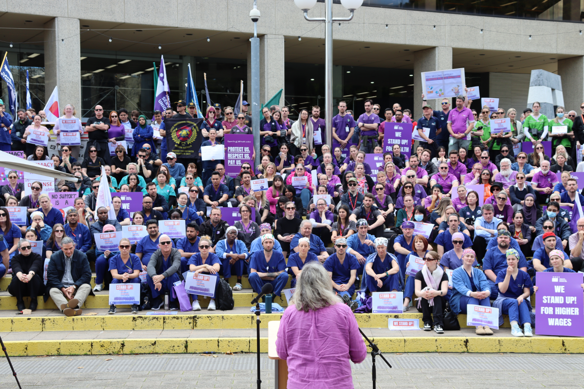 Big group of health workers listening to someone speak at a lectern in front of them while gathered on stairs.