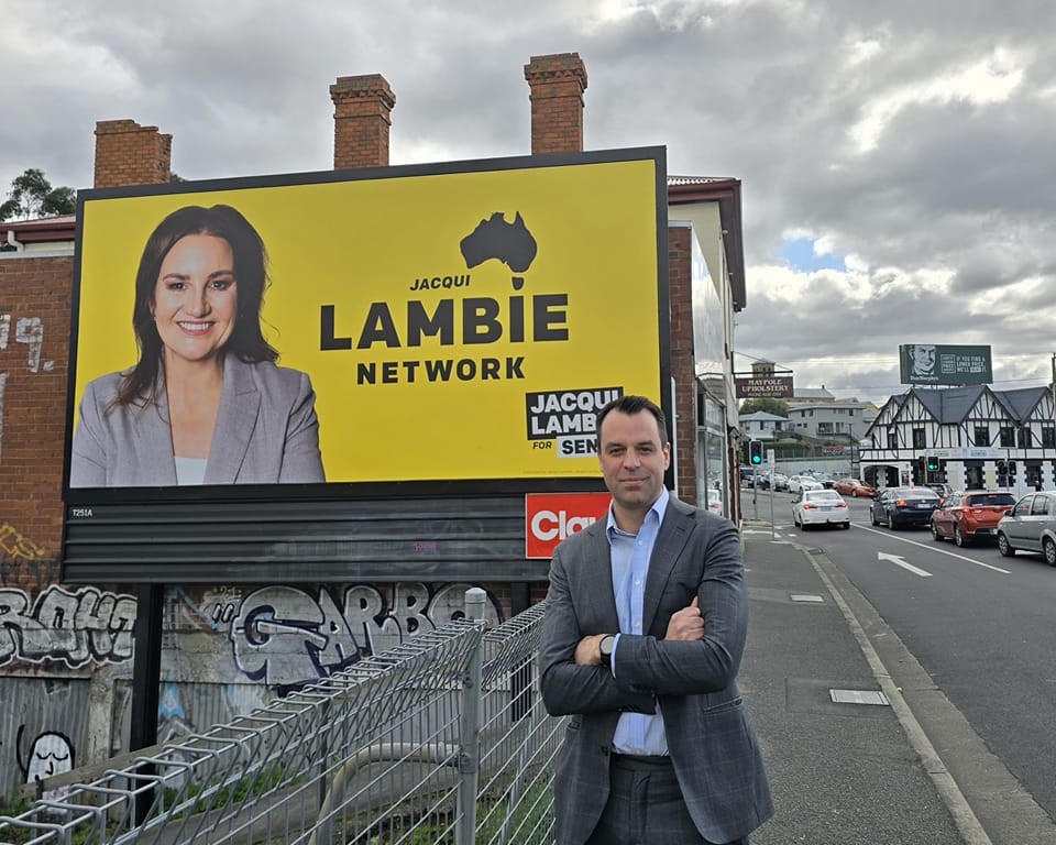 Josh Willie standing in front of a Jacqui Lambie Network sign next to a main road.