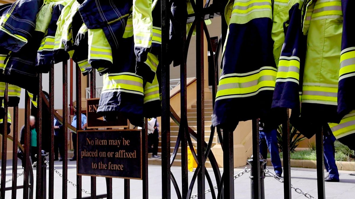 Firefighter jackets hanging off a fence outside NSW Parliament, with a sign reading not to hang anything on it.