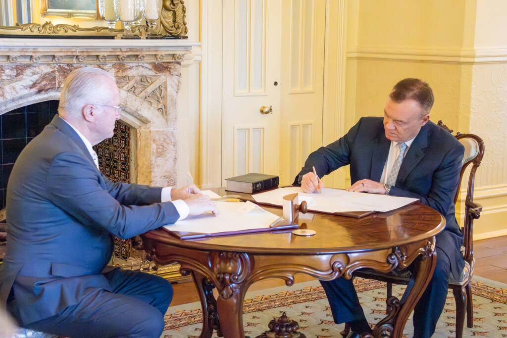 two men sitting at a table, with one signing a document