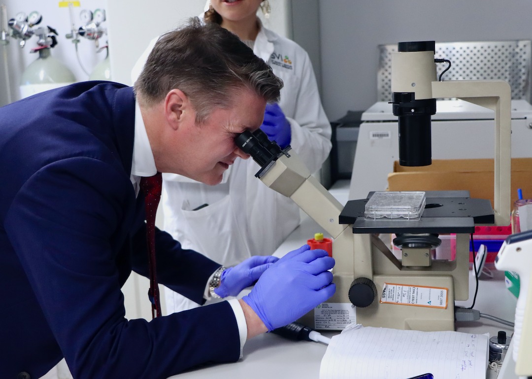 A man in a suit using a microscope with a lab technician behind him