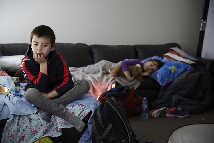 A young boy looking despondent, with his sister lying nearby on a couch and their belongings around them