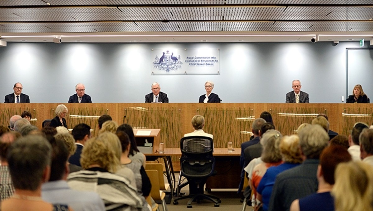 A group of commissioners sitting on a lateral table, while fronting a group of seated journalists and lawyers.