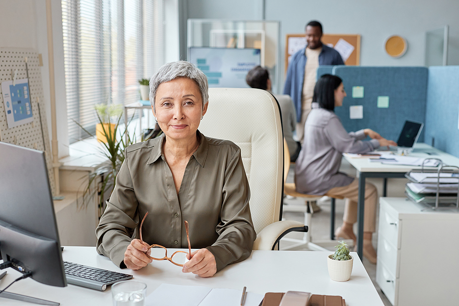 Front-view portrait of elegant senior businesswoman working at desk in open office setting and smiling at camera