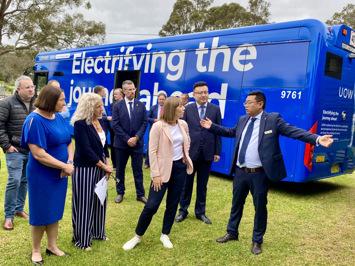 A man addressing a group of people standing on a lawn near a battery electric bus